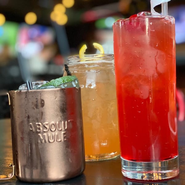 Three cocktails on a bar: a copper mug, a mason jar, and a tall red drink.
