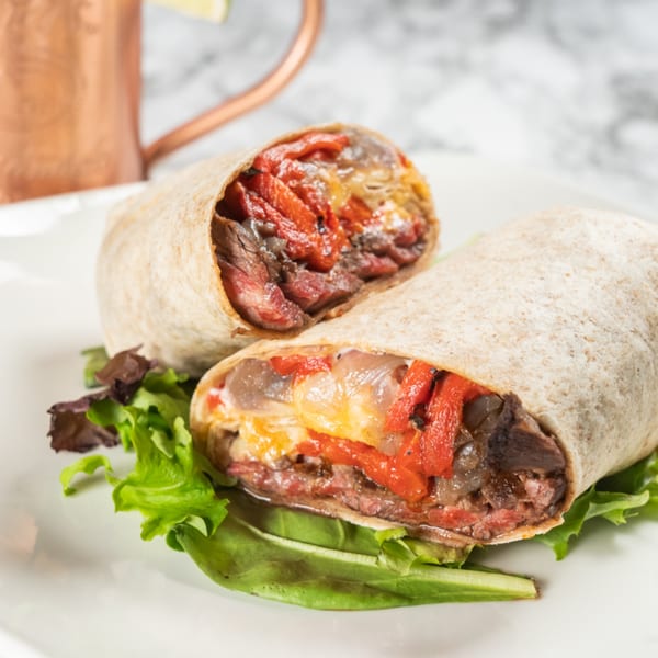 Two halves of a steak and pepper wrap on a white plate with lettuce, a copper mug in the background.