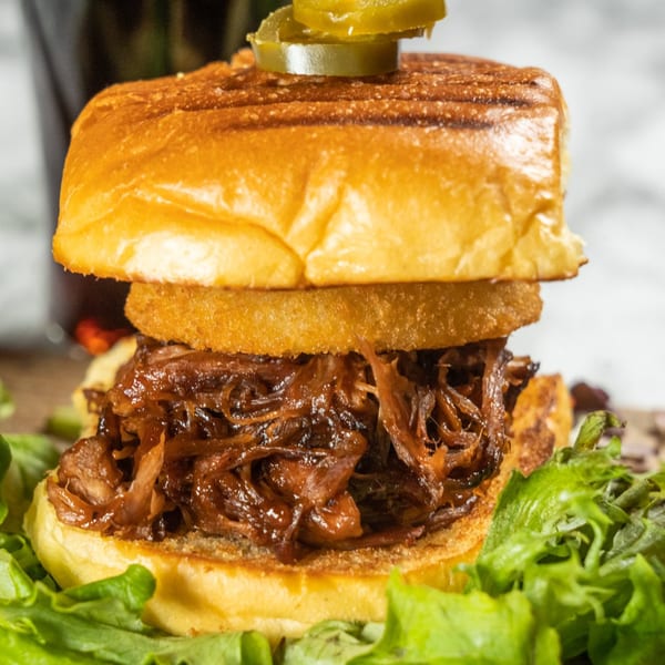 Close-up of a pulled pork sandwich with an onion ring, lettuce, and jalapeños on a brioche bun.