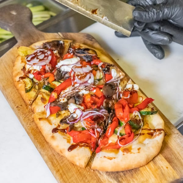A chef in black gloves slices a flatbread pizza topped with vegetables and cheese on a wooden board.