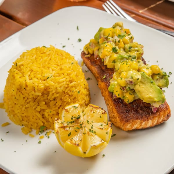 American salmon dish with mango avocado salsa, yellow rice, and a grilled lemon on a white plate.