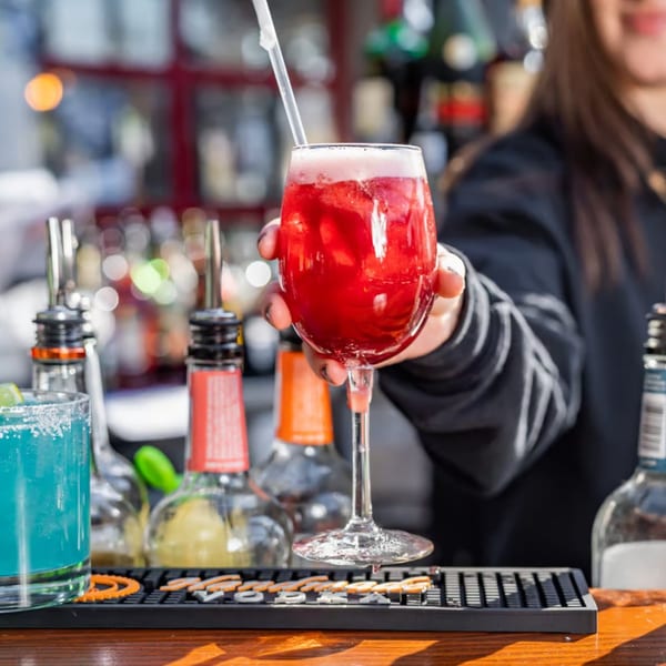 A bartender's hand holds up a red cocktail with a straw, with other drinks and bottles blurred in the background.