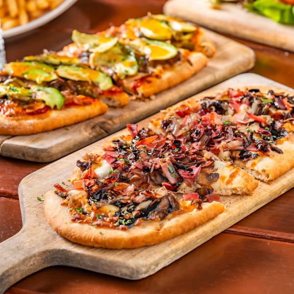 Two American flatbread pizzas on wooden boards, one with avocado, the other with bacon and mushrooms.